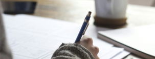 person writing on brown wooden table near white ceramic mug